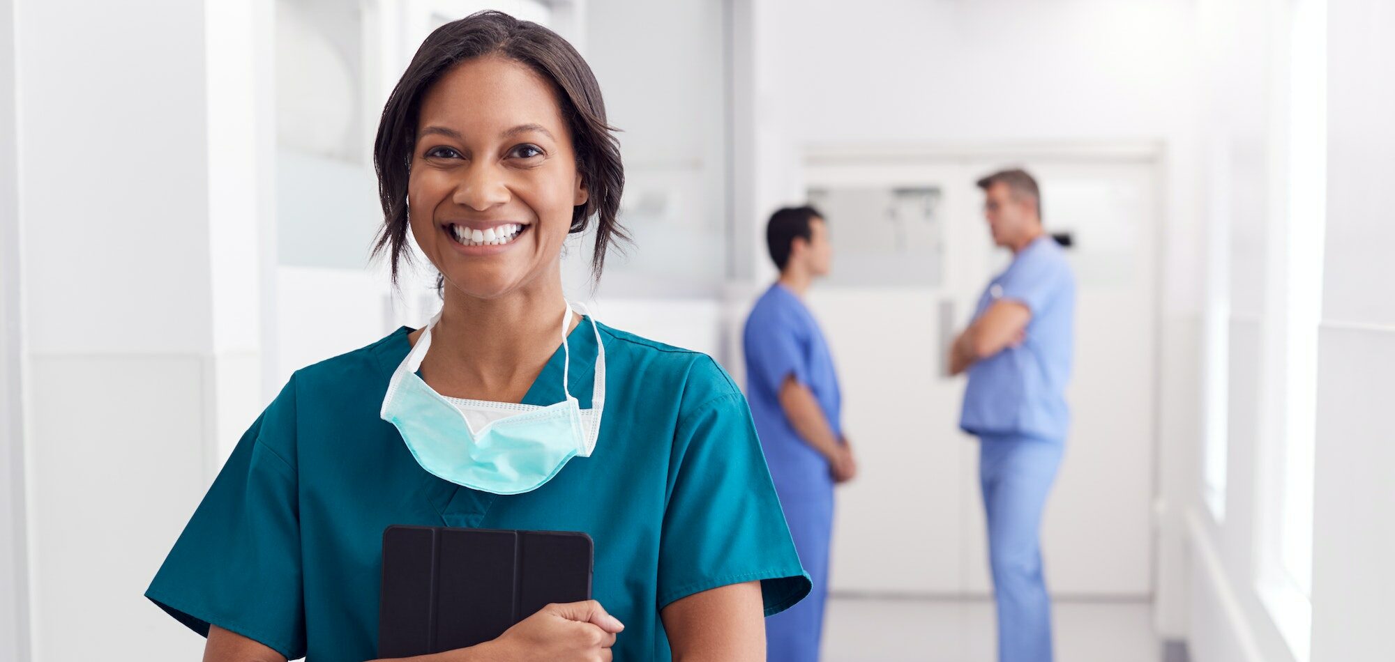 Portrait Of Smiling Female Doctor Wearing Scrubs In Hospital Corridor Holding Digital Tablet